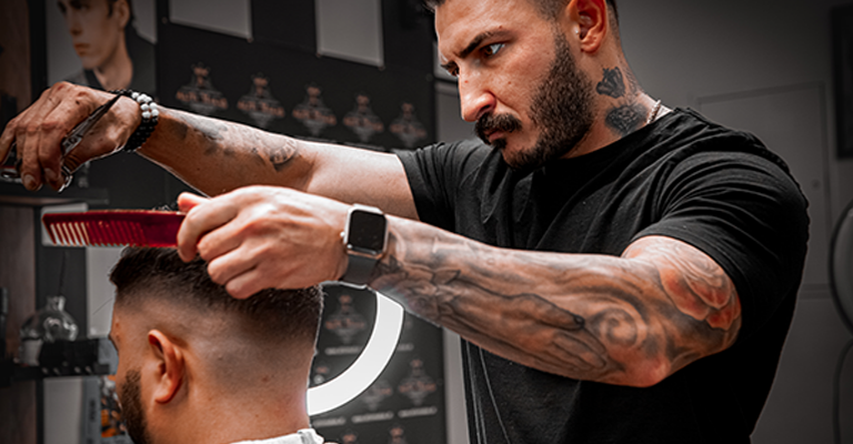 Barber mit tätowiertem Arm schneidet konzentriert mit Kamm und Schere die Haare eines Kunden